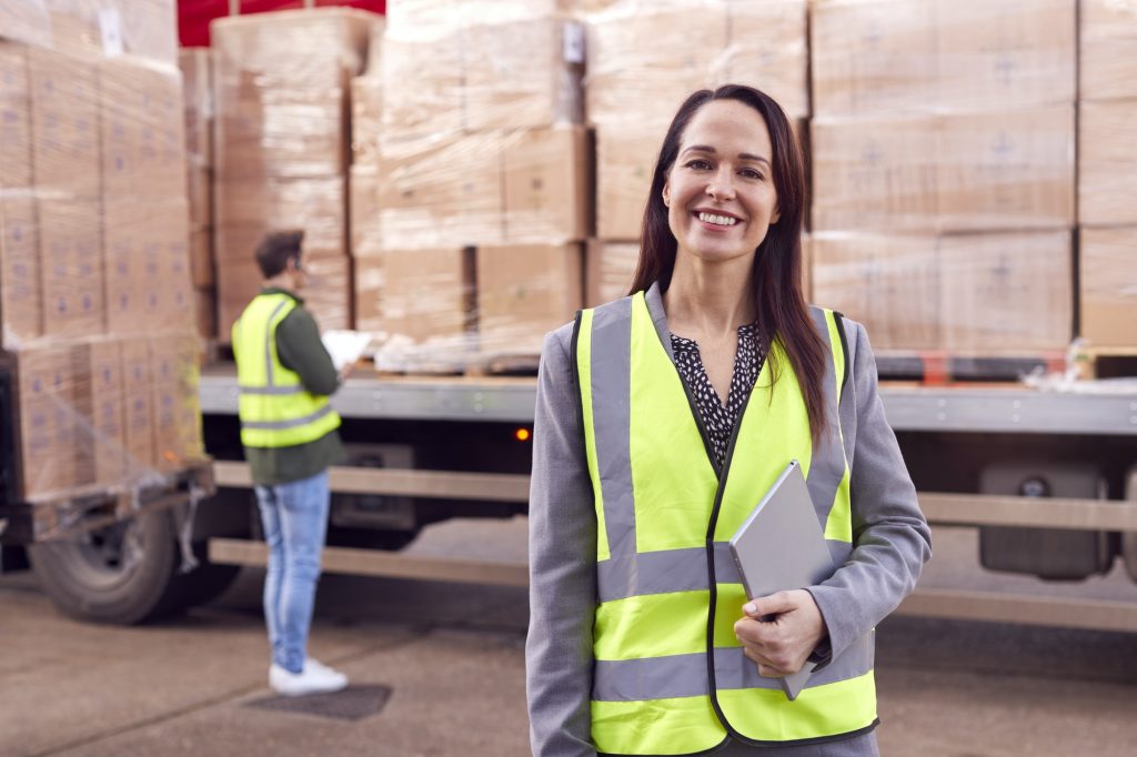 Portrait Of Female Freight Haulage Manager Standing By Truck Being Loaded By Fork Lift